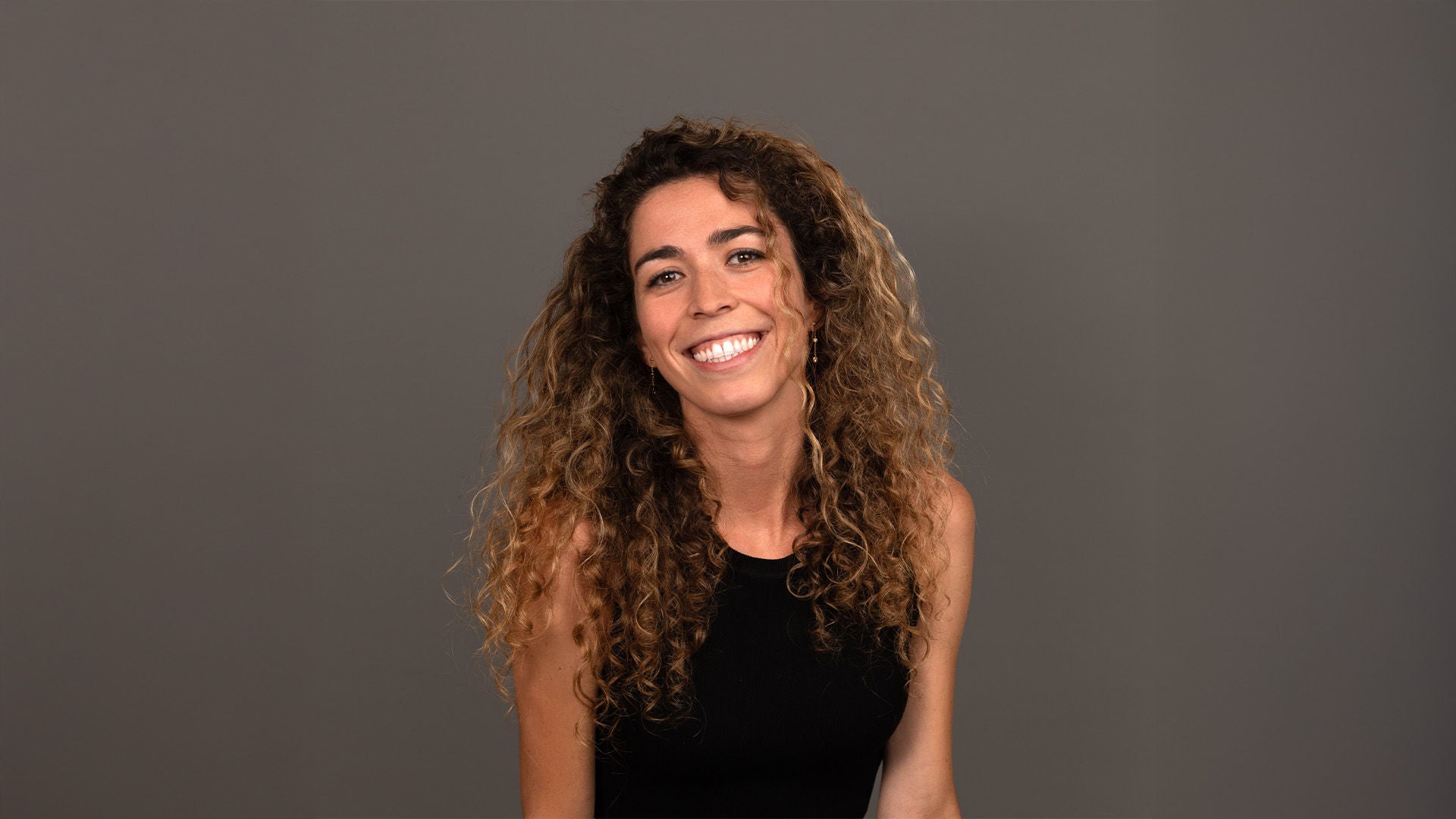 Frau mit langen lockigen Haaren im schwarzen ärmellosen Oberteil, lächelnd, vor grauem Hintergrund.