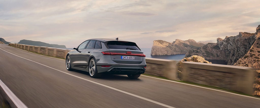 Ein grauer Audi A6 Avant fährt auf einer kurvigen Küstenstraße mit beeindruckender Felslandschaft und Meerblick im Hintergrund. Die Szenerie wird von einer warmen Abendstimmung begleitet.