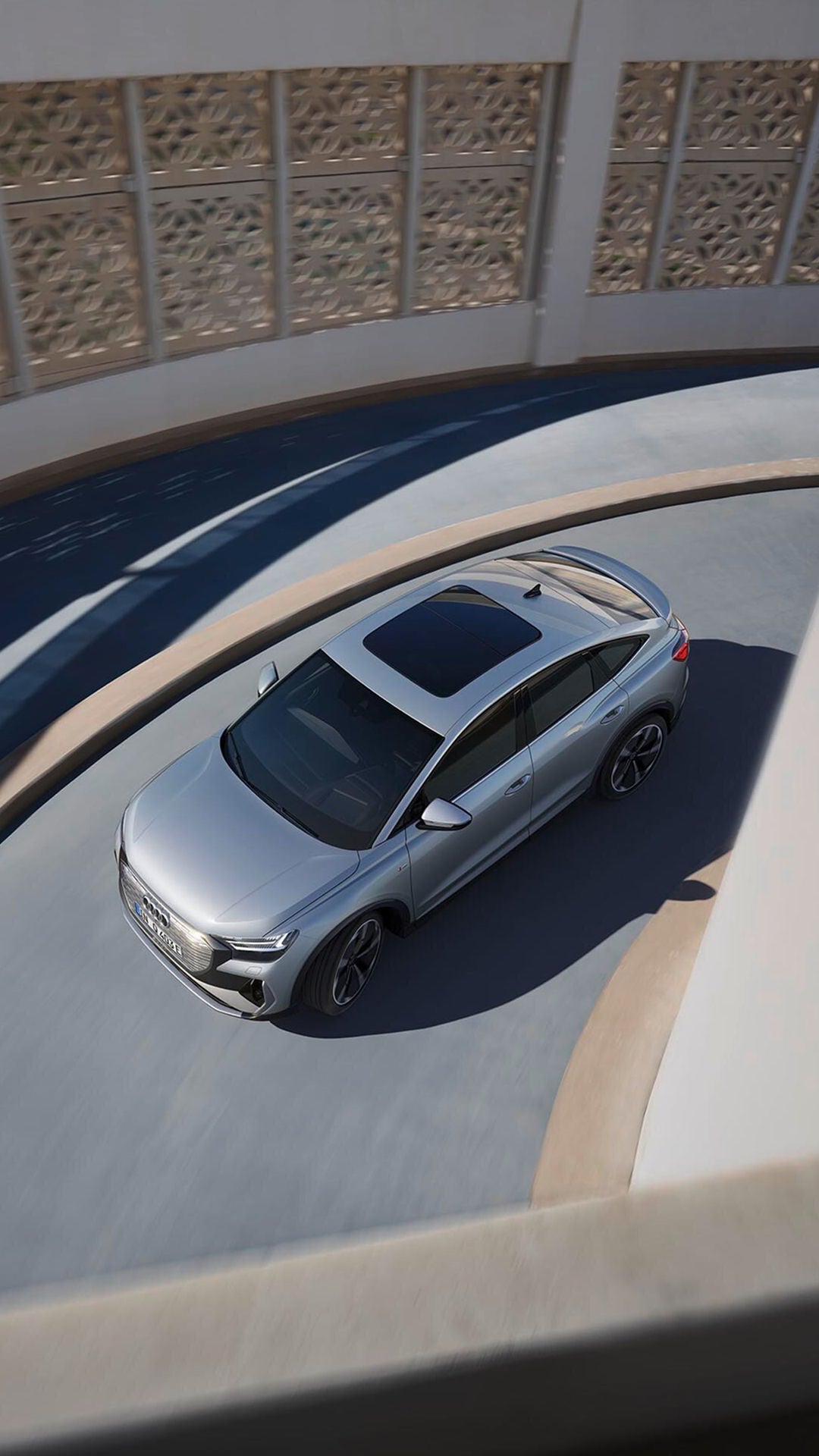 A silver Audi Q4 Sportback e-tron vehicle is captured from above, elegantly manoeuvring along a curved pathway in a modern environment.