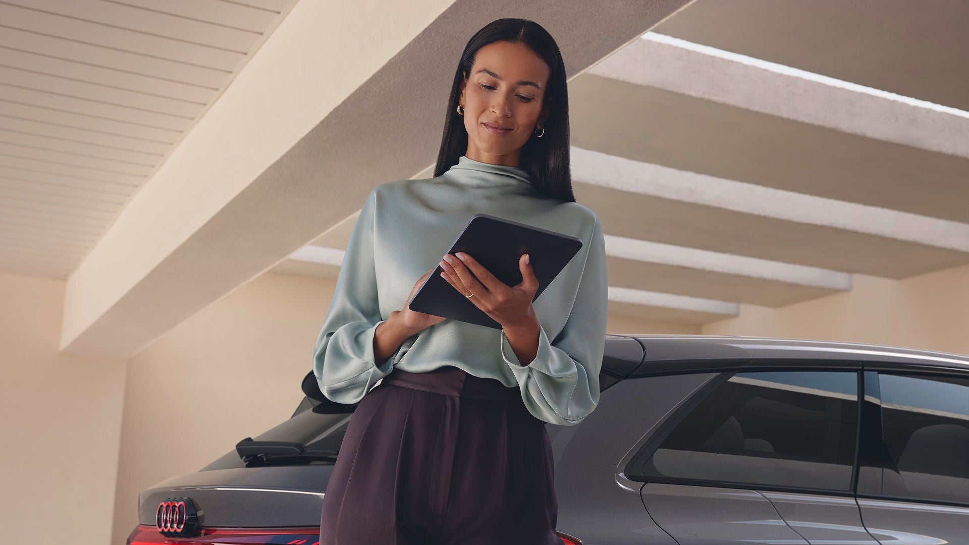 A woman wearing a light blue blouse stands under a modern carport, looking at a tablet. A grey car is parked nearby. The scene is calm and focused.