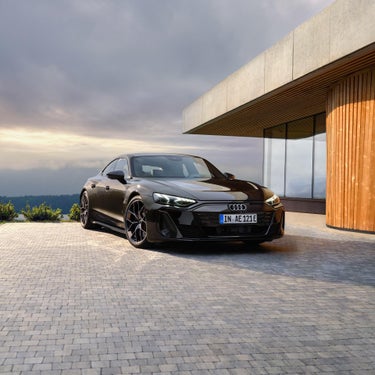 Ein schwarzer Audi S e-tron GT als Audi exclusive edition steht vor einem modernen Haus mit Holz- und Betonelementen auf einer gepflasterten Auffahrt. Der Himmel ist teils bewölkt und erzeugt eine dramatische Atmosphäre.