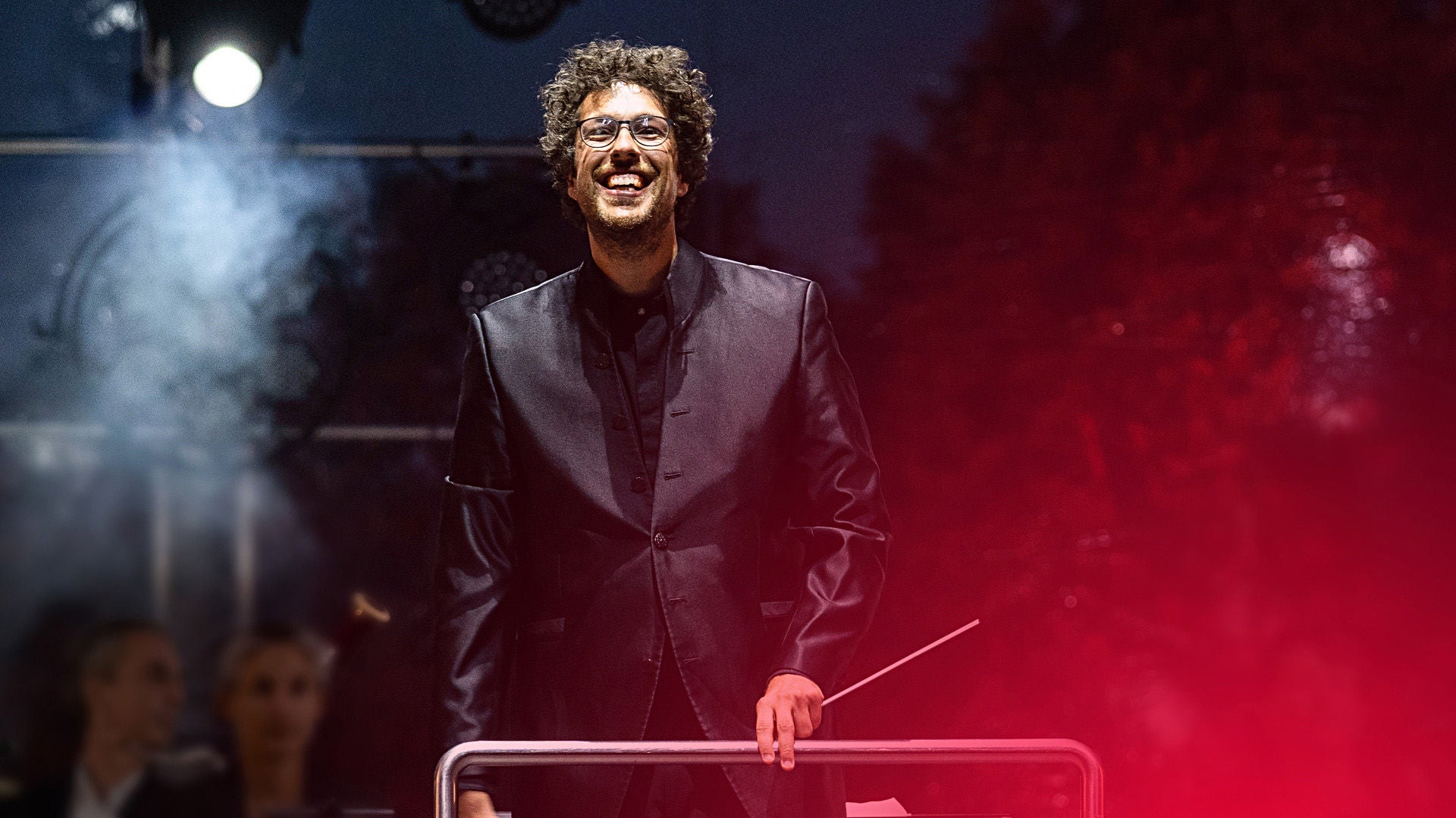 Dirigent auf Konzertbühne im Abendlicht von Scheinwerfern angestrahlt.