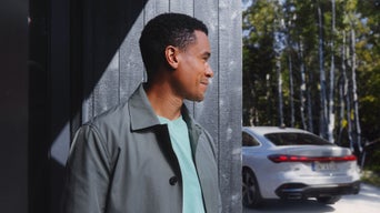 Un homme souriant se tient debout devant un mur sombre, vêtu d'un manteau gris et d'un t-shirt vert. À l'arrière-plan, une Audi A5 blanche est garée devant une forêt de bouleaux baignée de soleil.