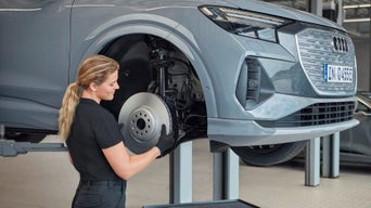Frau prüft Bremsscheibe an grauem Audi auf Hebebühne in heller Werkstatt.