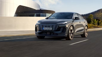 Frontansicht eines dunklen Audi Q6 Sportback e-tron, der auf einer Straße fährt. Im Hintergrund sieht man ein modernes Gebäude mit markanter Architektur und einen grünen Hügel.