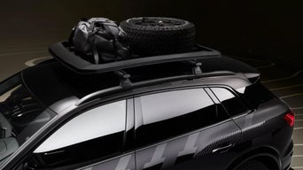 Schwarzer Audi mit Dachkorb, auf dem ein Ersatzrad und eine Sporttasche befestigt sind, vor dunklem Hintergrund.