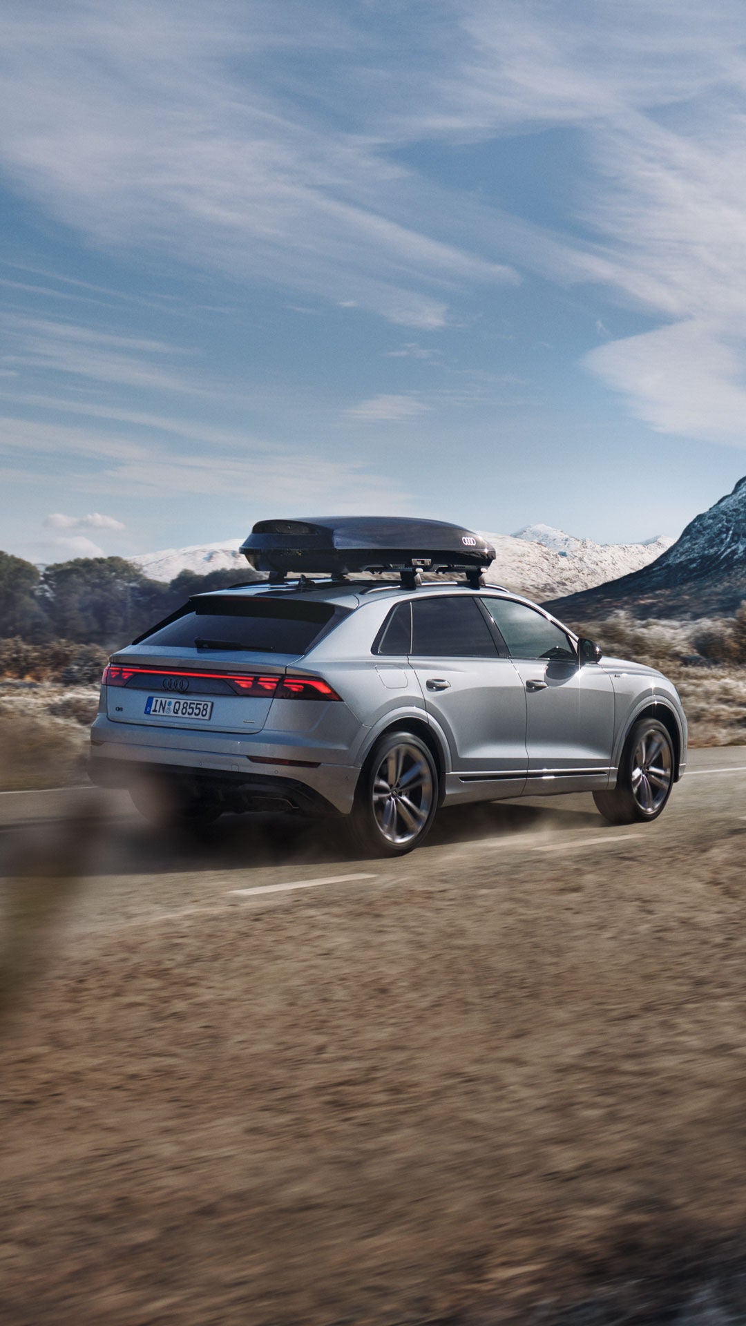 Ein silberner Audi mit Dachbox fährt in einer hügeligen Wüstenlandschaft mit Schnee auf den Bergen