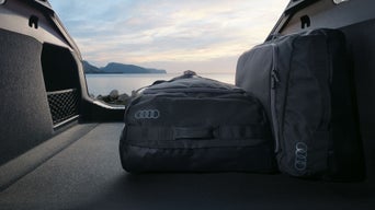 Zwei schwarze Audi-Reisetaschen stehen im offenen Kofferraum eines Autos mit Blick auf Meer und Berge bei Sonnenuntergang.