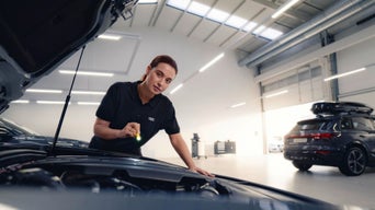 Eine Frau mit einem Tablet in der Hand in einem Autohaus im Hintergrund steht ein Audi und zwei Leute mit einem Laptop.