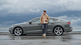Ein Mann lehnt mit einem Getränk an einem grauen Auto, bei bewölktem Himmel auf nasser Straße nahe dem Wasser.