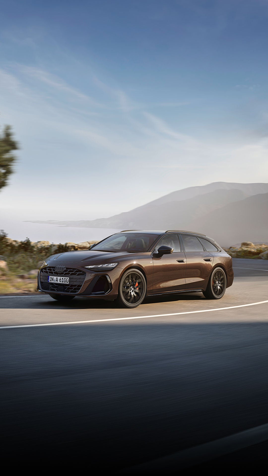 A brown A6 Avant driving in a scenic route with tree and mountains in the background