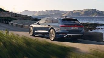 Ein blauer Audi S6 Avant e-tron dynamisch fährt auf einer Küstenstraße, im Hintergrund Berge und Meer bei Sonnenuntergang.