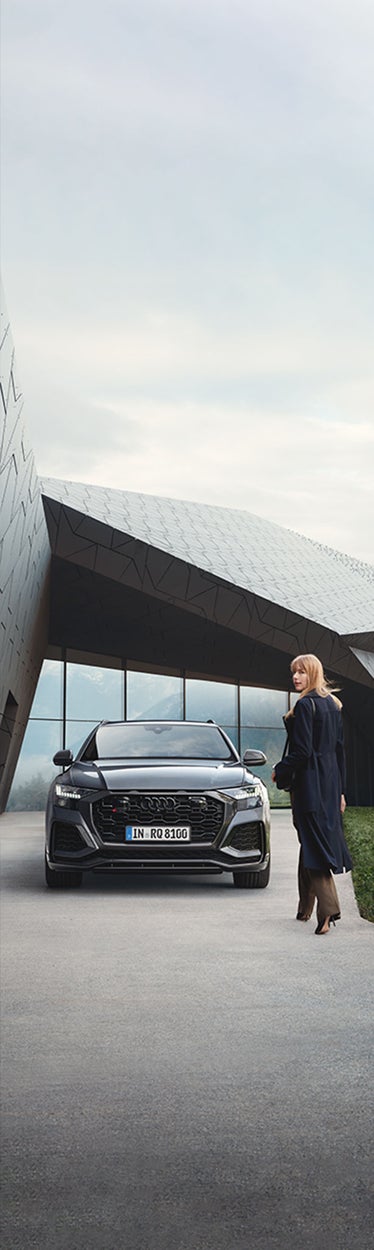 Frau steht vor schwarzem Audi-SUV an modernem Gebäude in den Bergen, Wolken und Landschaft im Hintergrund.