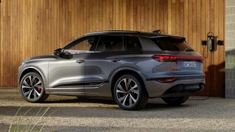 Seitenprofil des SQ6 SUV e-tron, welcher vor einer Holzhausfassade steht und an eine an der Hauswand montierte Ladestation angeschlossen ist.