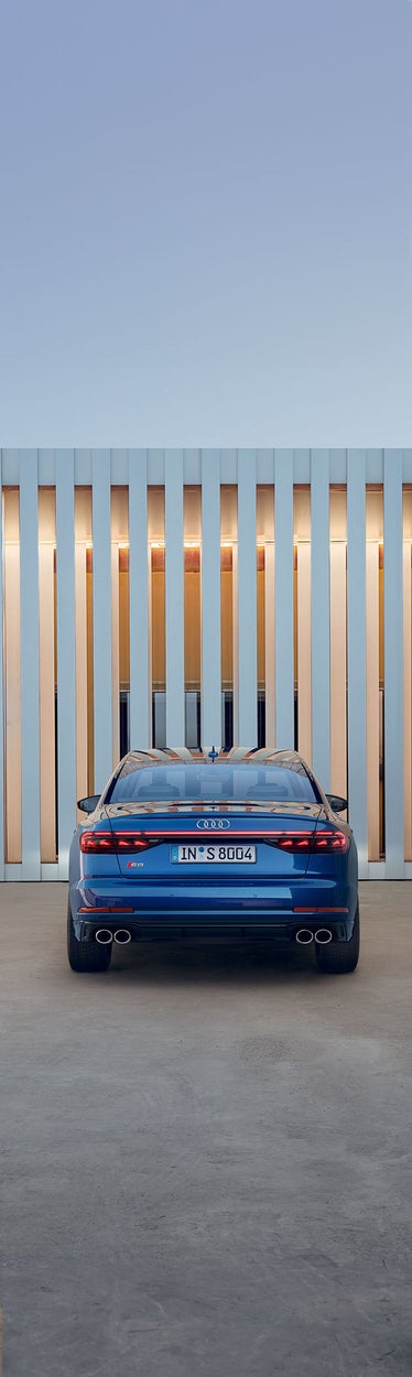 Heckansicht eines blauen Audi S8 TFSI mit vier Auspuffrohren vor einer hellen Fassade mit vertikalen Lichtstreifen.