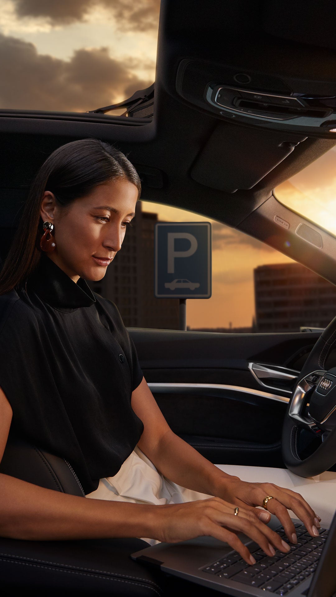 Frau arbeitet bei Sonnenuntergang mit Laptop auf dem Fahrersitz eines geparkten Audi, im Hintergrund ein Parkschild.