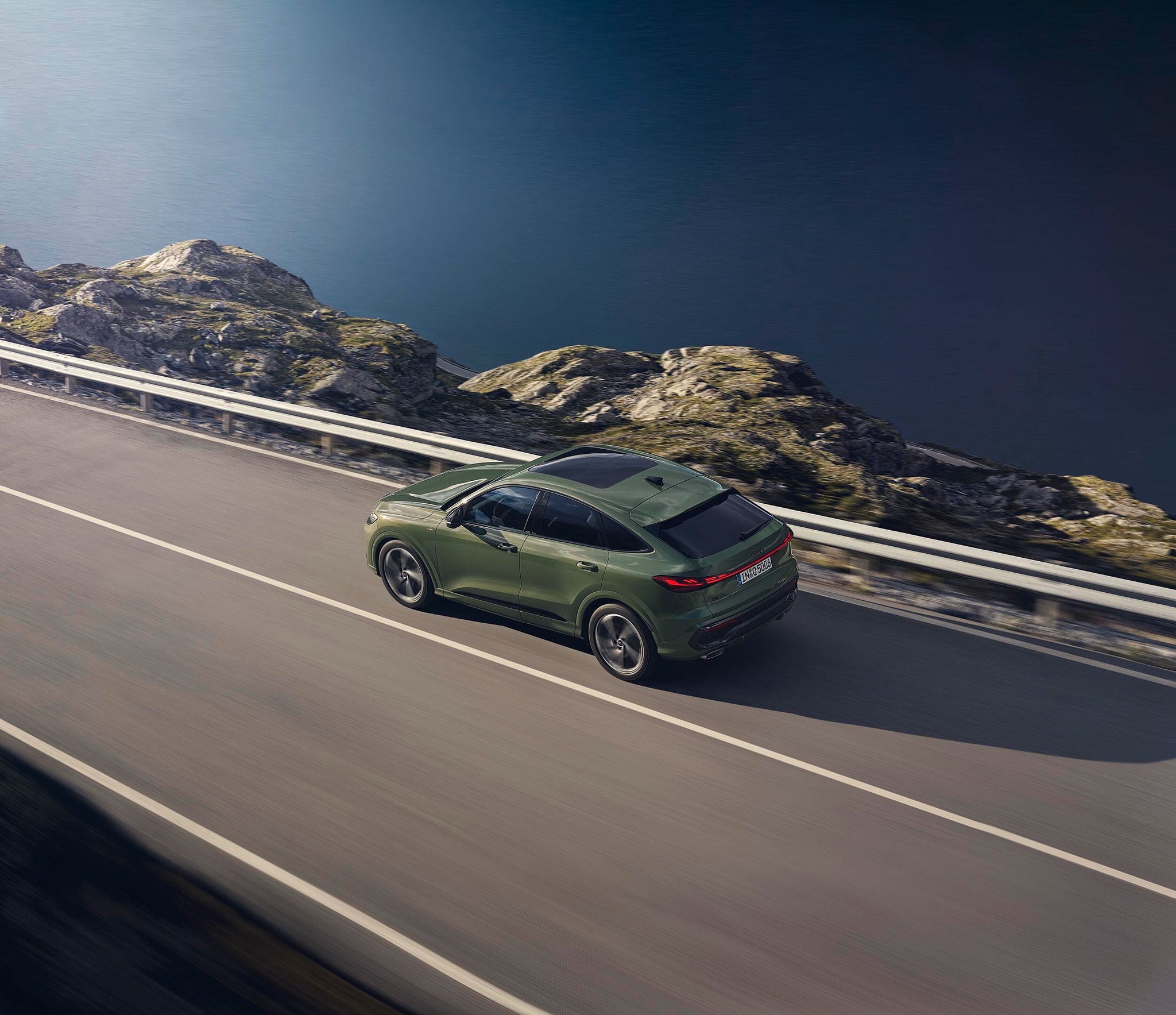 Grüner Audi Q5 fährt auf Küstenstraße entlang eines Gewässers, felsige Landschaft und Leitplanke am Straßenrand.