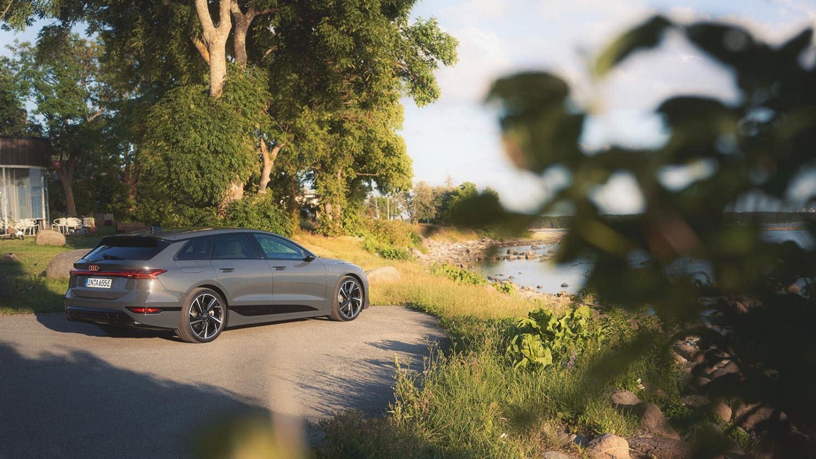 Grå Audi A6 e-tron kombi fotograferad snett bakifrån, parkerad på en grusväg vid vattnet i kvällsljus, omgiven av grönska och träd.