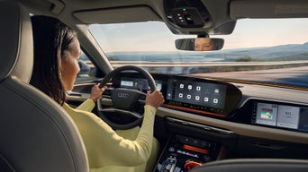 Close-up van de cockpit van een Audi, schuin van achter de bestuurdersstoel genomen.  Aan het stuur zit een vrouw.  Ze kijkt naar een kustlandschap.  Haar ogen zijn te zien in de achteruitkijkspiegel.