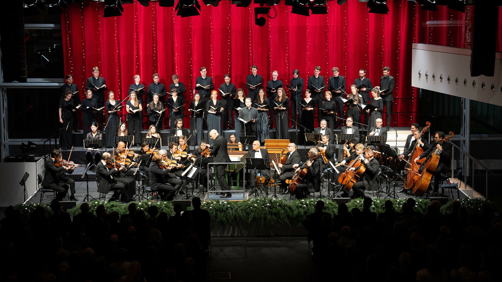 Dirigent, Orchester und Chor führen live vor Publikum auf einer Bühne mit rotem Vorhang klassische Musik auf.
