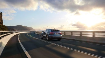 Ein Audi Q6 e-tron fährt bei Sonnenuntergang auf einer Küstenstraße mit Geländer, rechts das Meer, links Berge