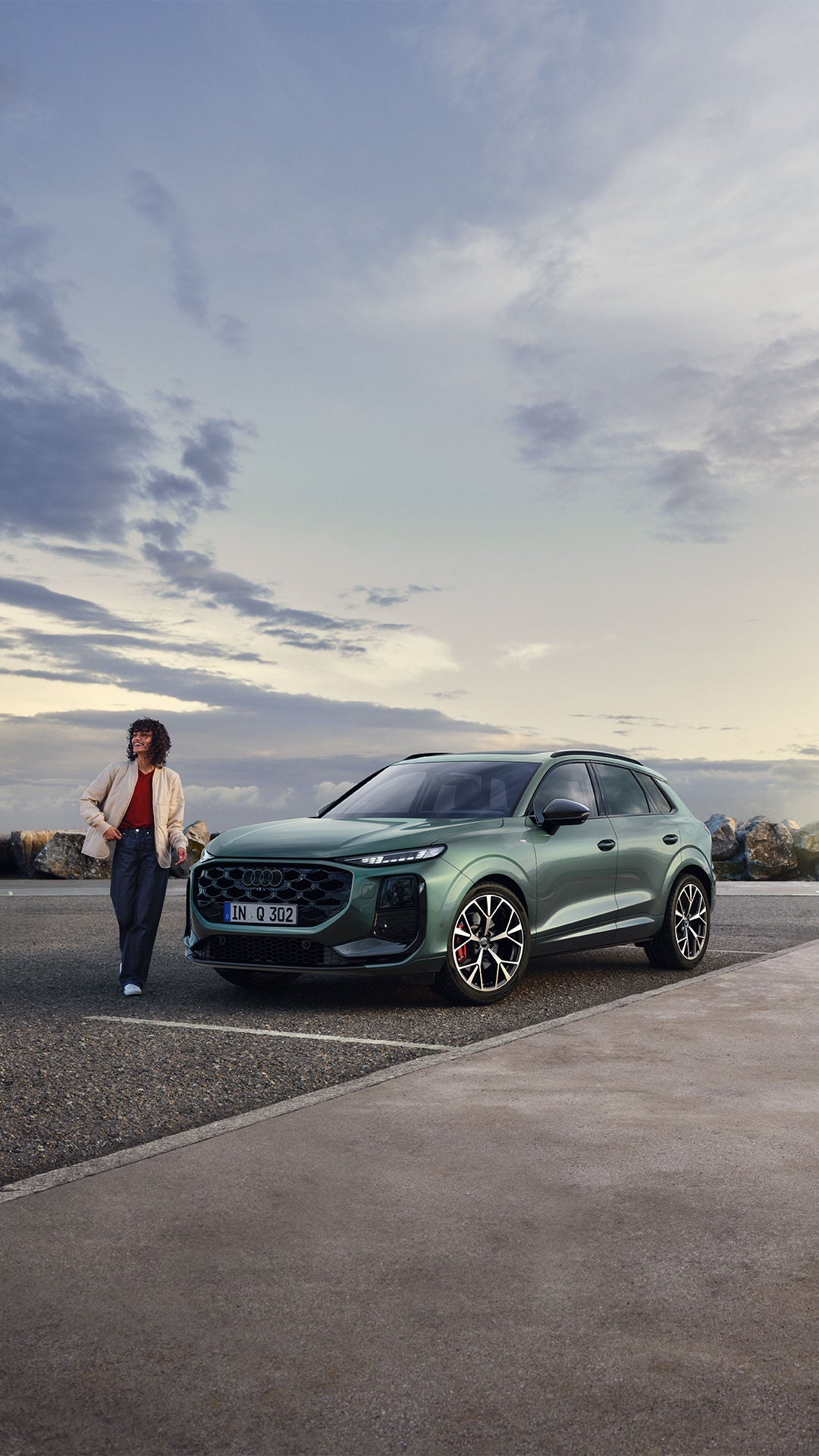 Audi Q3 Sportback vert foncé stationné, avec une personne souriante qui marche à côté, sur un terrain goudronné près de rochers et sous un ciel nuageux.