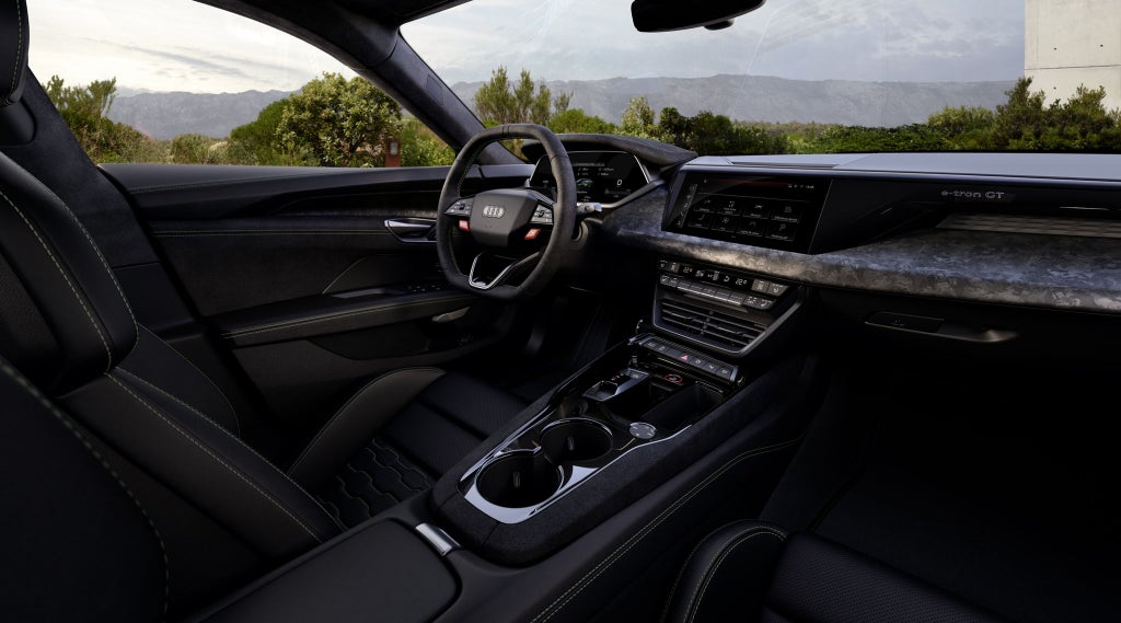 Interior de um Audi RS e-tron GT performance com couro preto, volante, tela sensível ao toque e vista para a paisagem campestre verdejante com montanhas.