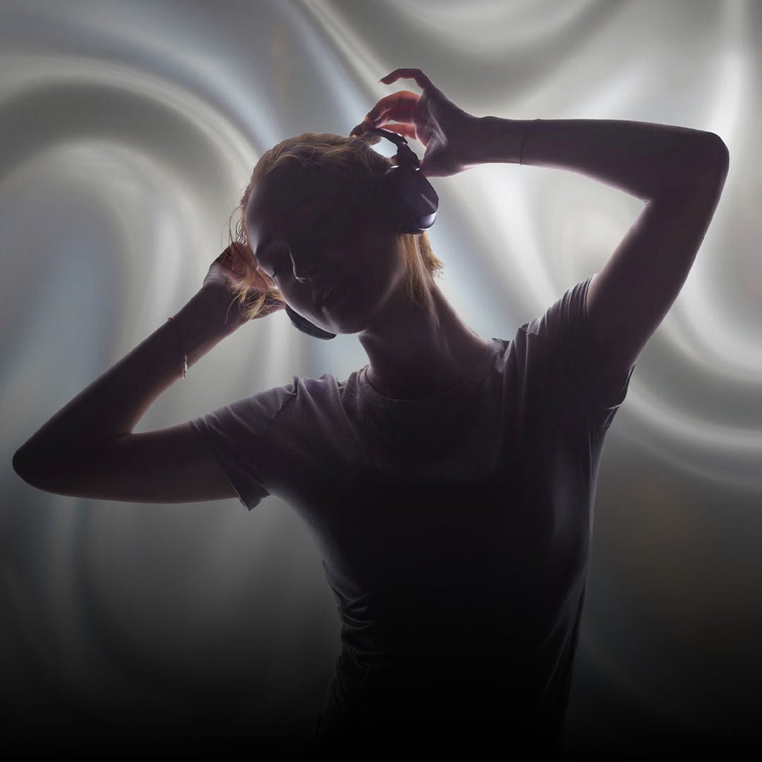 Person mit Kopfhörern im Profil hebt die Hände an den Kopfhörer, vor hellem, weich verschwommenem Hintergrund.