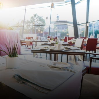 Heller Restaurantbereich mit gedeckten Tischen, weißen Tischdecken, Pflanzen und roten Stühlen am Fenster.