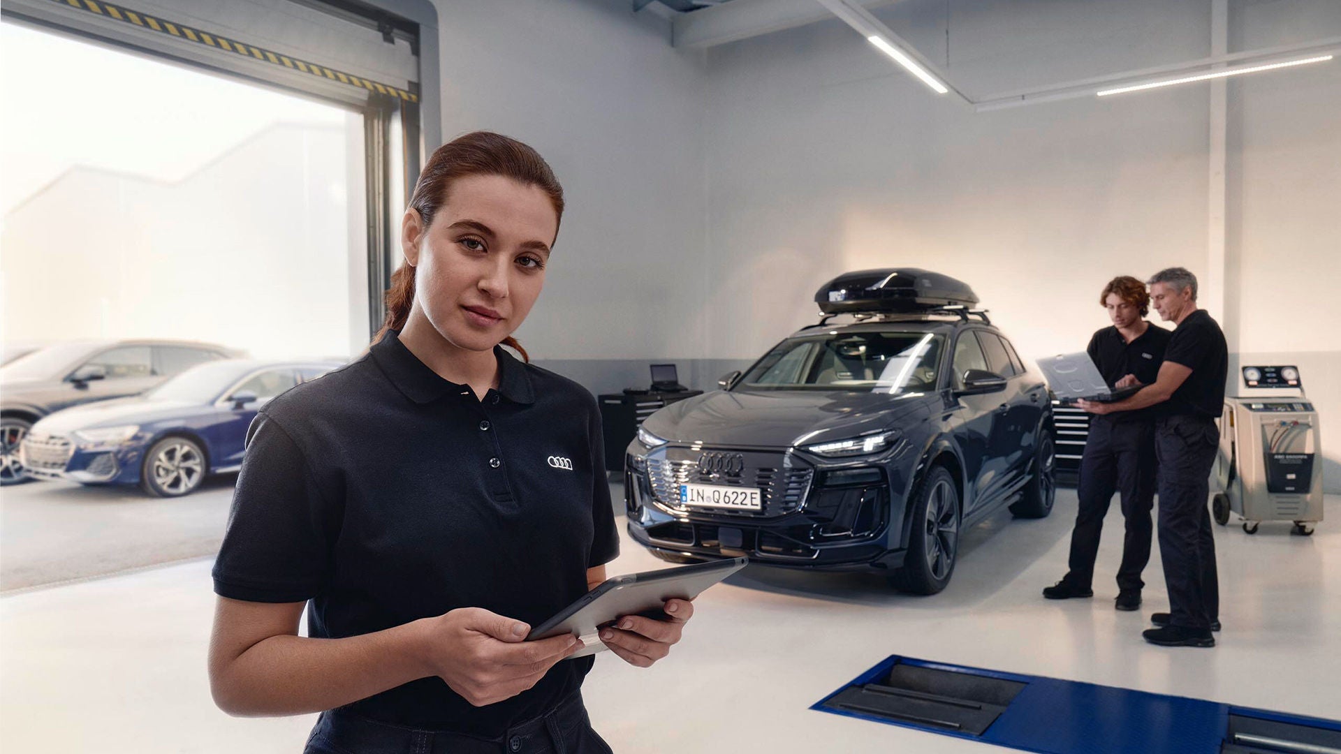Person mit Tablet in einer Audi Werkstatt, mit einem Elektrofahrzeug im Servicebereich im Hintergrund.