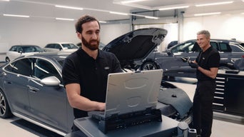 Mechaniker in schwarzer Audi-Uniform prüft Fahrzeug mit Laptop in einer Werkstatt.