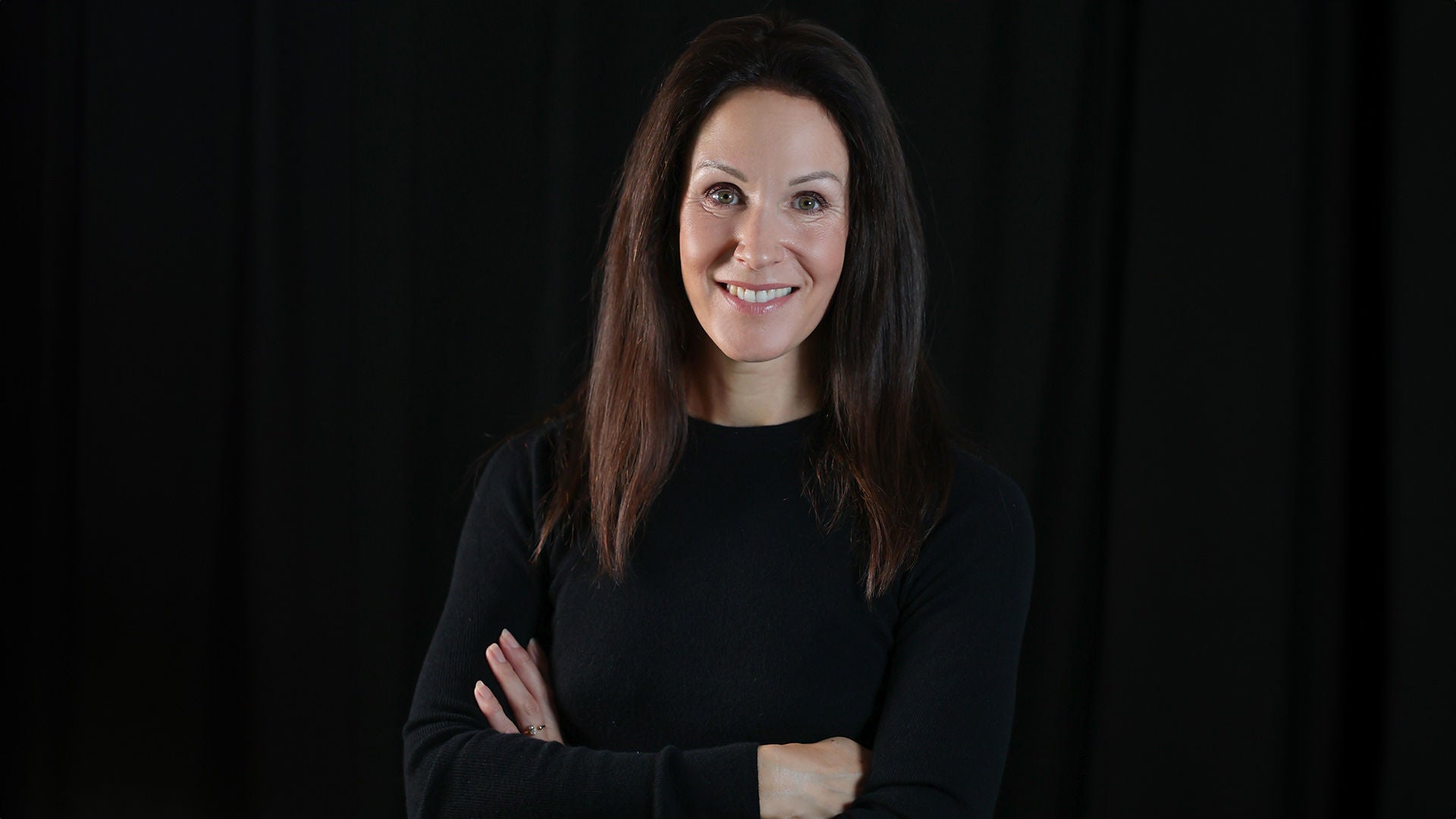 Lächelnde Frau mit langen dunklen Haaren, Arme verschränkt, schwarzes Oberteil vor dunklem Hintergrund.
