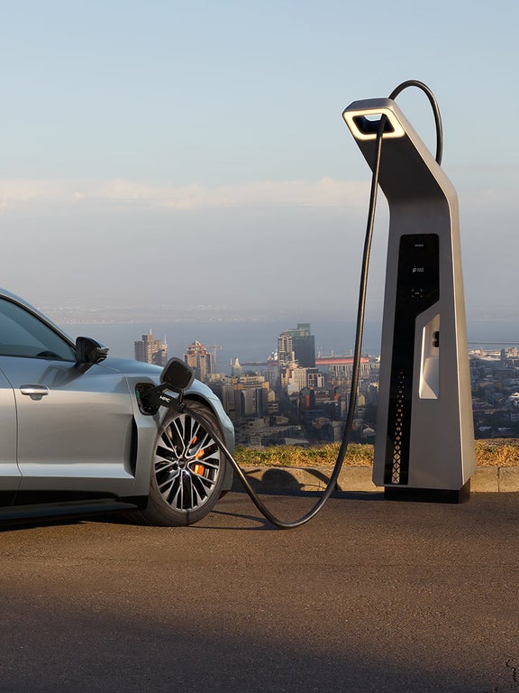 Ein silberner Audi S e-tron GT lädt an einer Ladestation mit Blick auf eine Stadt. Ein Mann im Anzug geht neben dem Auto entlang.