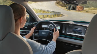 Close-up van de cockpit van een Audi, schuin van achter de bestuurdersstoel genomen.  Aan het stuur zit een vrouw.  Ze kijkt naar een kronkelige weg.  Haar ogen zijn te zien in de achteruitkijkspiegel.