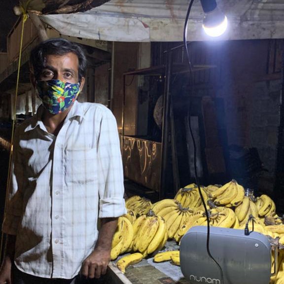 Man with mask leaning with his right hand on a market table with bananas. Looks into the camera.