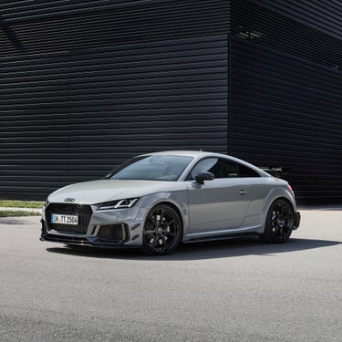 Grauer Audi TT RS mit schwarzem Heckspoiler steht auf Asphalt vor einer schwarzen Fassade, fotografiert leicht schräg von vorn.