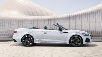 Weißes Audi Cabriolet mit geöffnetem Verdeck vor moderner, wellenförmiger Fassade mit glatter Kachelstruktur.