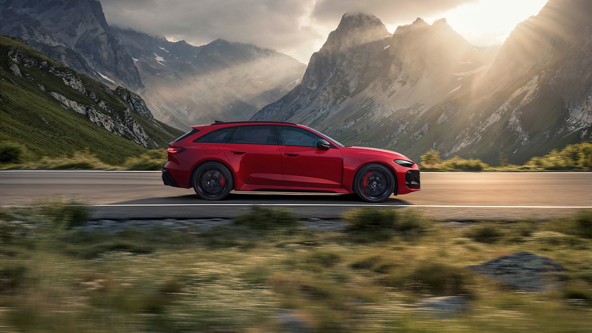 Roter Audi RS 5 Avant fährt vor Bergkulisse im Gegenlicht; Sonnenstrahlen und felsige Gipfel im Hintergrund.