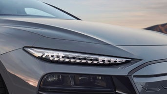 Close-up of a sleek gray car's front with modern LED headlights, showcasing a stylish design against a soft sunset backdrop.