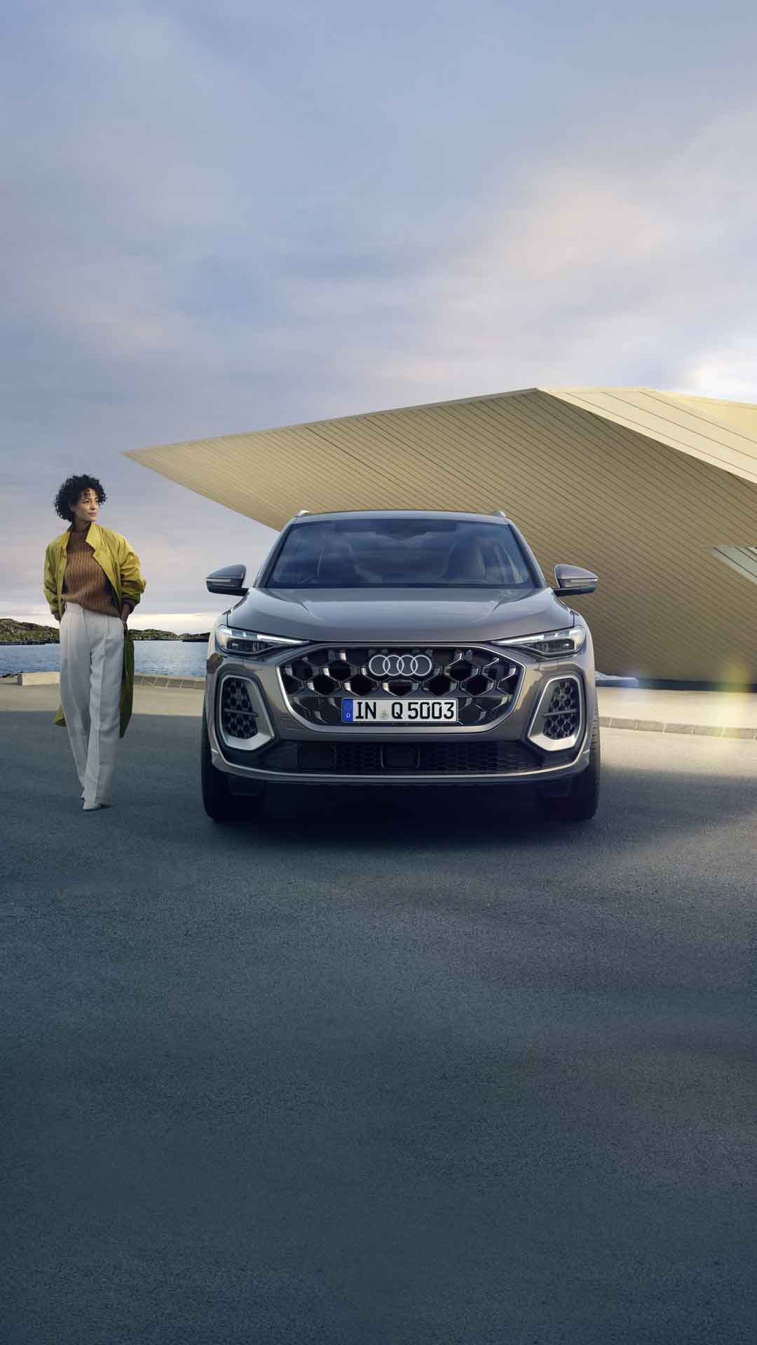 Der Q5 SUV im Frontprofil auf einem Parkplatz direkt an der Küste, dahinter ein kleines, futuristisches Haus. Neben dem Q5 SUV steht eine junge, modisch gekleidete Frau.	