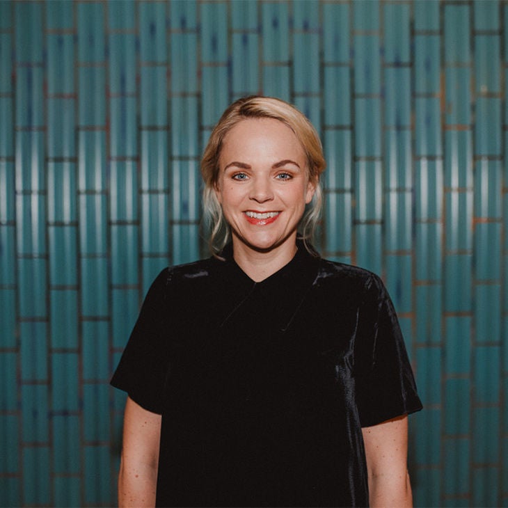 Lächelnde Frau mit blondem Haar und schwarzem Oberteil steht vor einer Wand aus türkisfarbenen, vertikalen Fliesen.