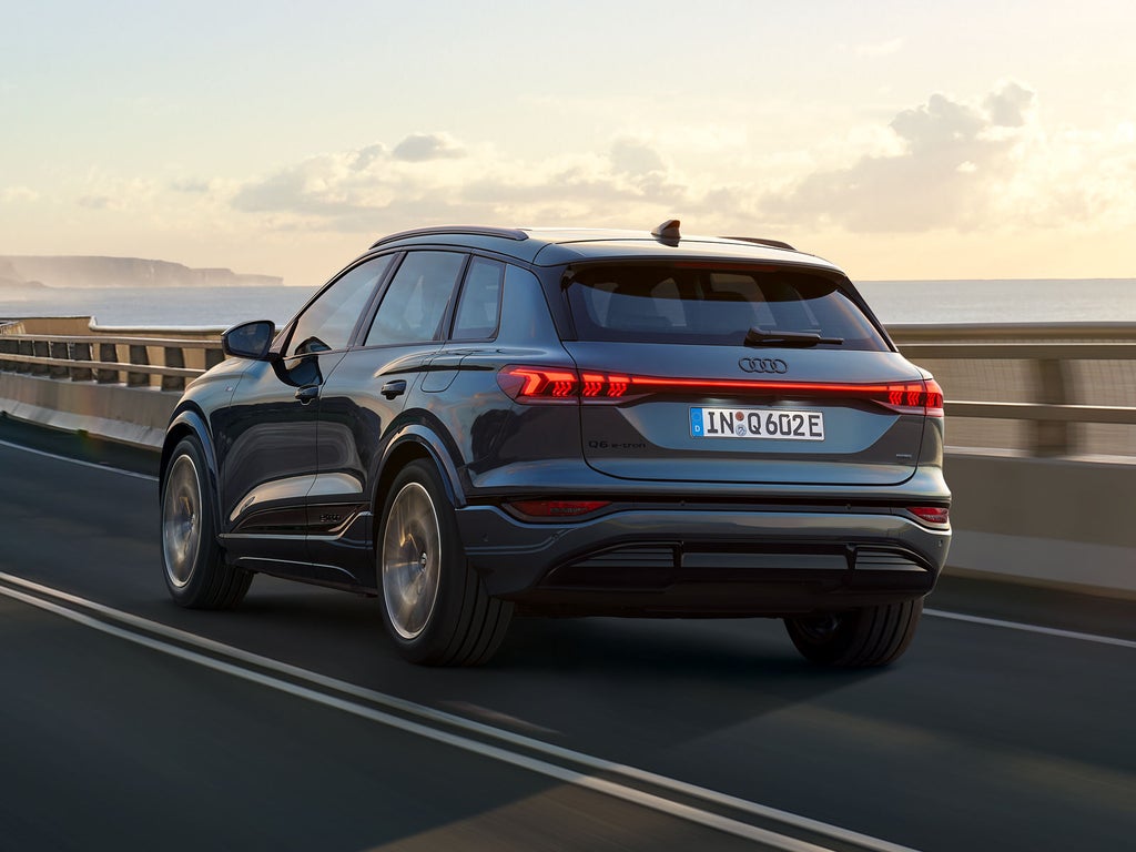 Schräge Heckaufnahme des Q6 SUV e-tron, welche über eine kurvige Straße am Meer fährt. Im Hintergrund sind Berge zu sehen.