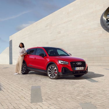 A person standing beside a red Audi SUV in front of a modern building.