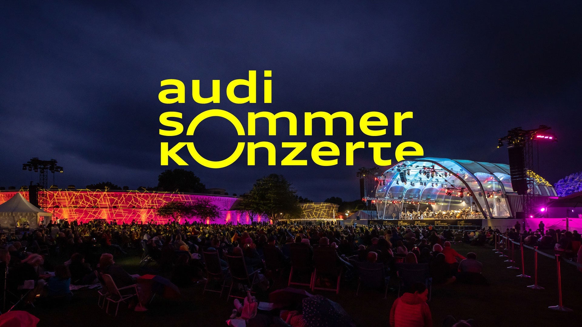 Hell beleuchtete Freiluftbühne am Abend, darüber gelber Schriftzug: Audi Sommerkonzerte.