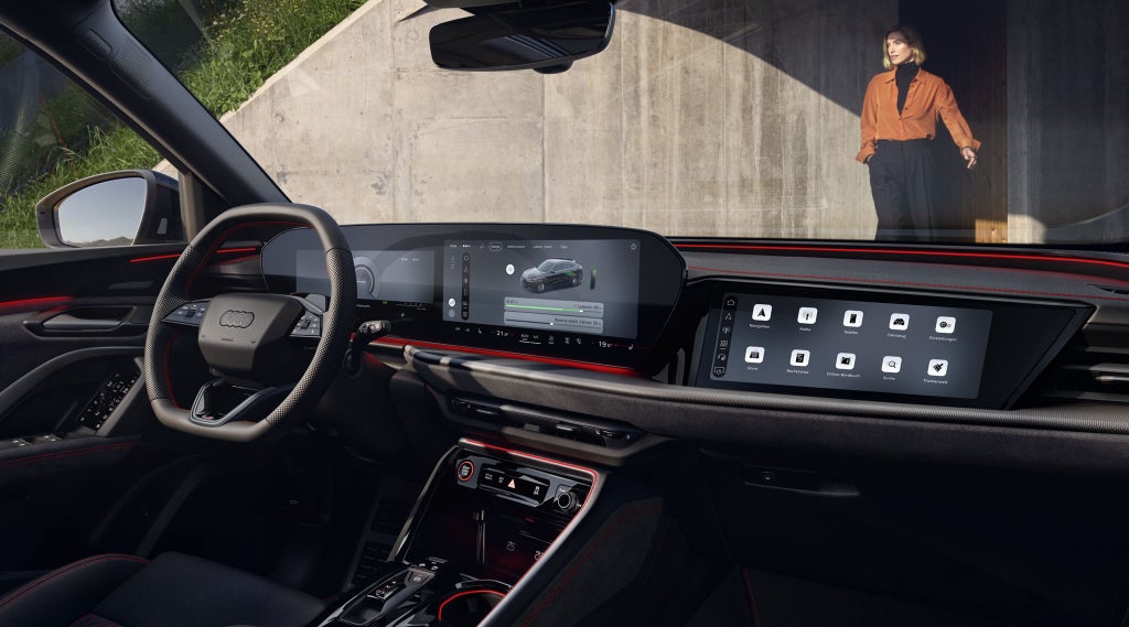 Der Innenraum des Q5 Sportback e-hybrid mit Fokus auf das Lenkrad und die digitalen Displays. Vor dem Fahrzeug steht eine modisch gekleidete, junge Frau.