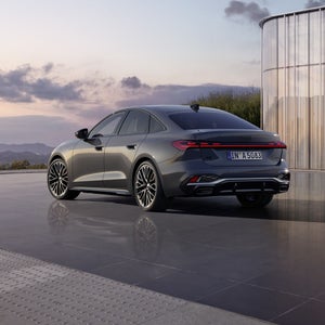 Die schwarze Edition One mit Akzenten in Magnesiumgrau der A5 Limousine in schrägen Heckprofil spiegelt sich in dem glatten Untergrund. Im Hintergrund sind Berge bei Dämmerung zu sehen.