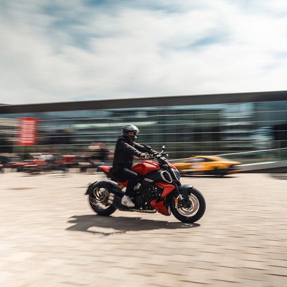 Motorradfahrer fährt dynamisch über Platz vor modernem Gebäude; unscharfer Hintergrund mit Autos und Passanten.