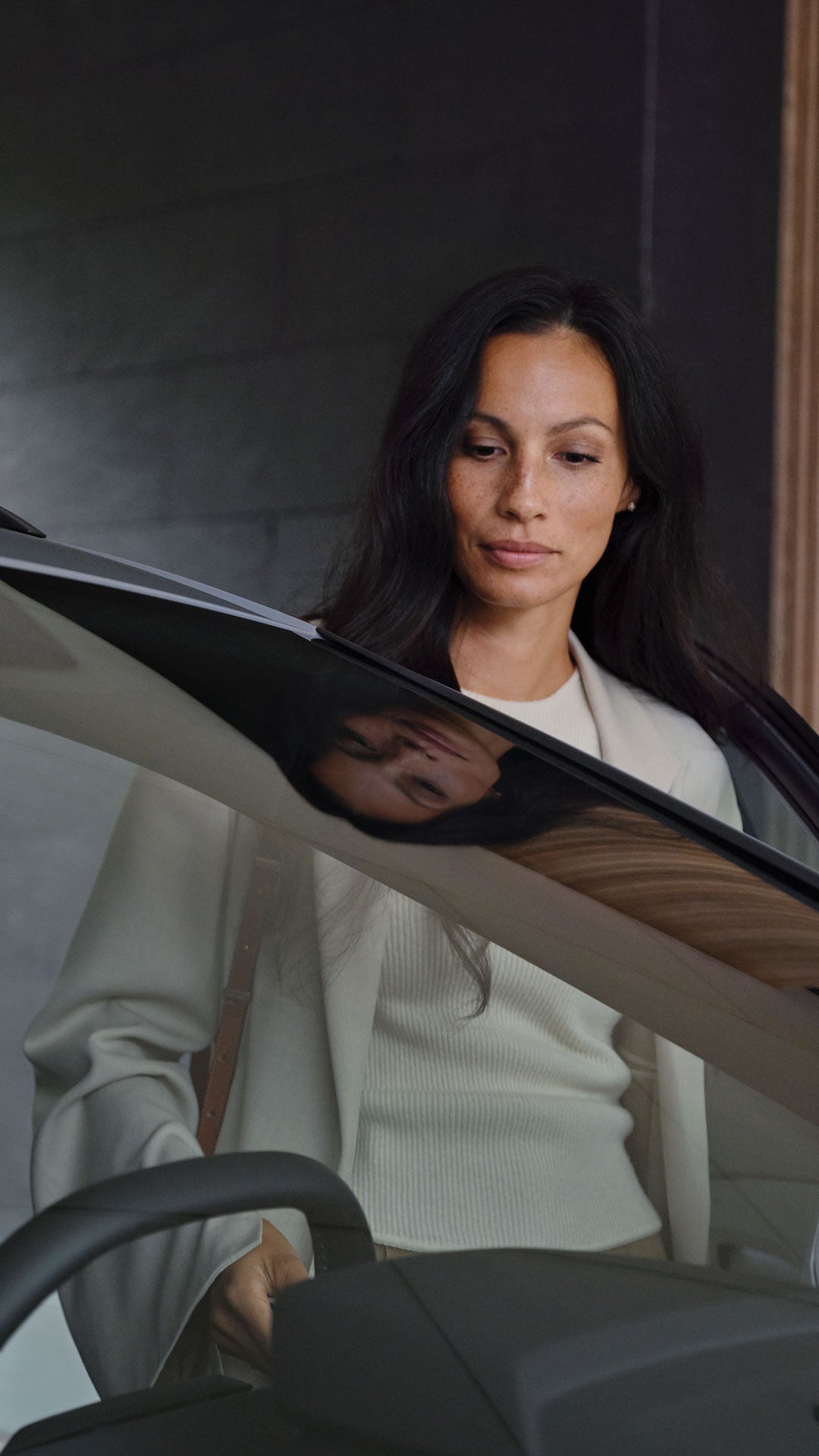 Frau mit langem dunklem Haar blickt durch die geöffnete Fahrertür eines Autos, ihr Spiegelbild ist in der Scheibe sichtbar.