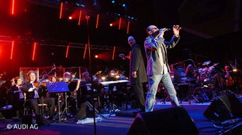 Mann steht auf einer Bühne inmitten eines Liveorchestras und singt