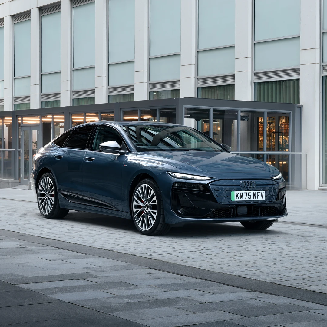 Dark blue Audi Sportback e-tron parked outside a modern office building, shown from the front three-quarter angle. The vehicle features a sleek aerodynamic design, LED headlights, large alloy wheels and a closed front grille. Floor-to-ceiling windows and illuminated interiors provide an urban backdrop under overcast daylight.